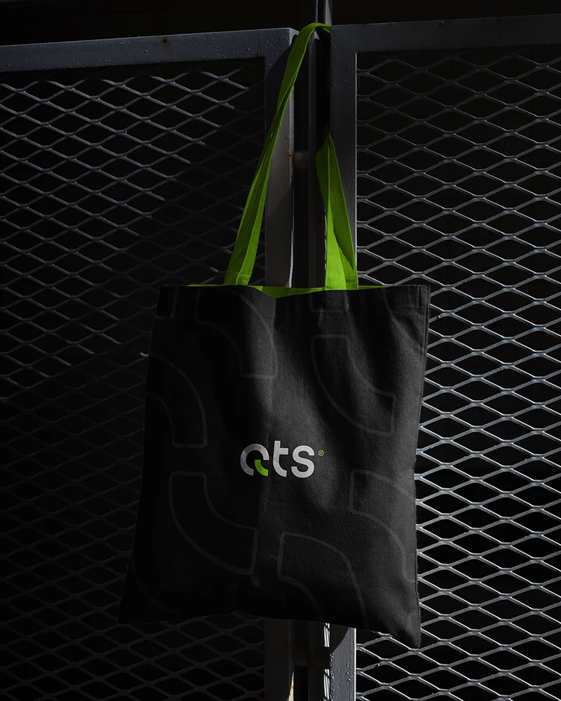 Black canvas tote bag featuring QTS logo on a metallic fence background